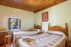 A room with two beds, a window with a view of greenery, a wooden ceiling, and a painting on the wall.