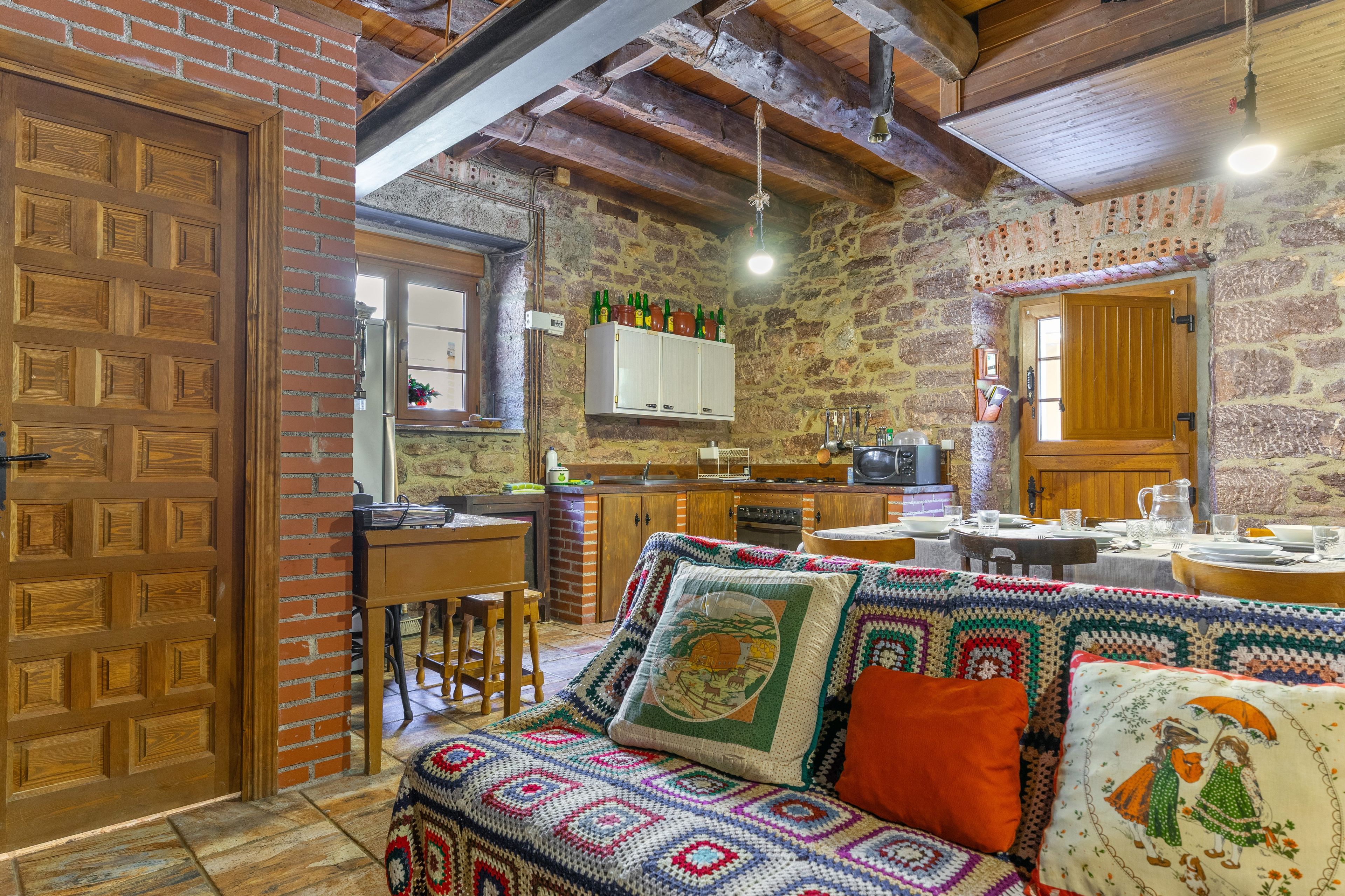 Un interior acogedor con una puerta de madera, un sofá estampado, una pared de piedra y una zona de cocina con armarios de madera y una lámpara colgante.