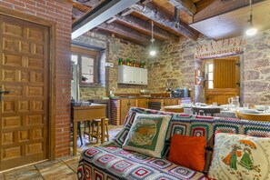 A cozy interior with a wooden door, a patterned sofa, a stone wall, and a kitchen area with wooden cabinets and a hanging light fixture.