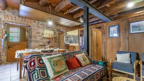 Una habitación acogedora con techo de madera, pared de piedra, una mesa de comedor para dos personas, un sofá colorido y un sillón azul.