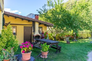 A rustic outdoor seating area with a wooden table and benches, surrounded by potted plants and a yellow building with a chimney.