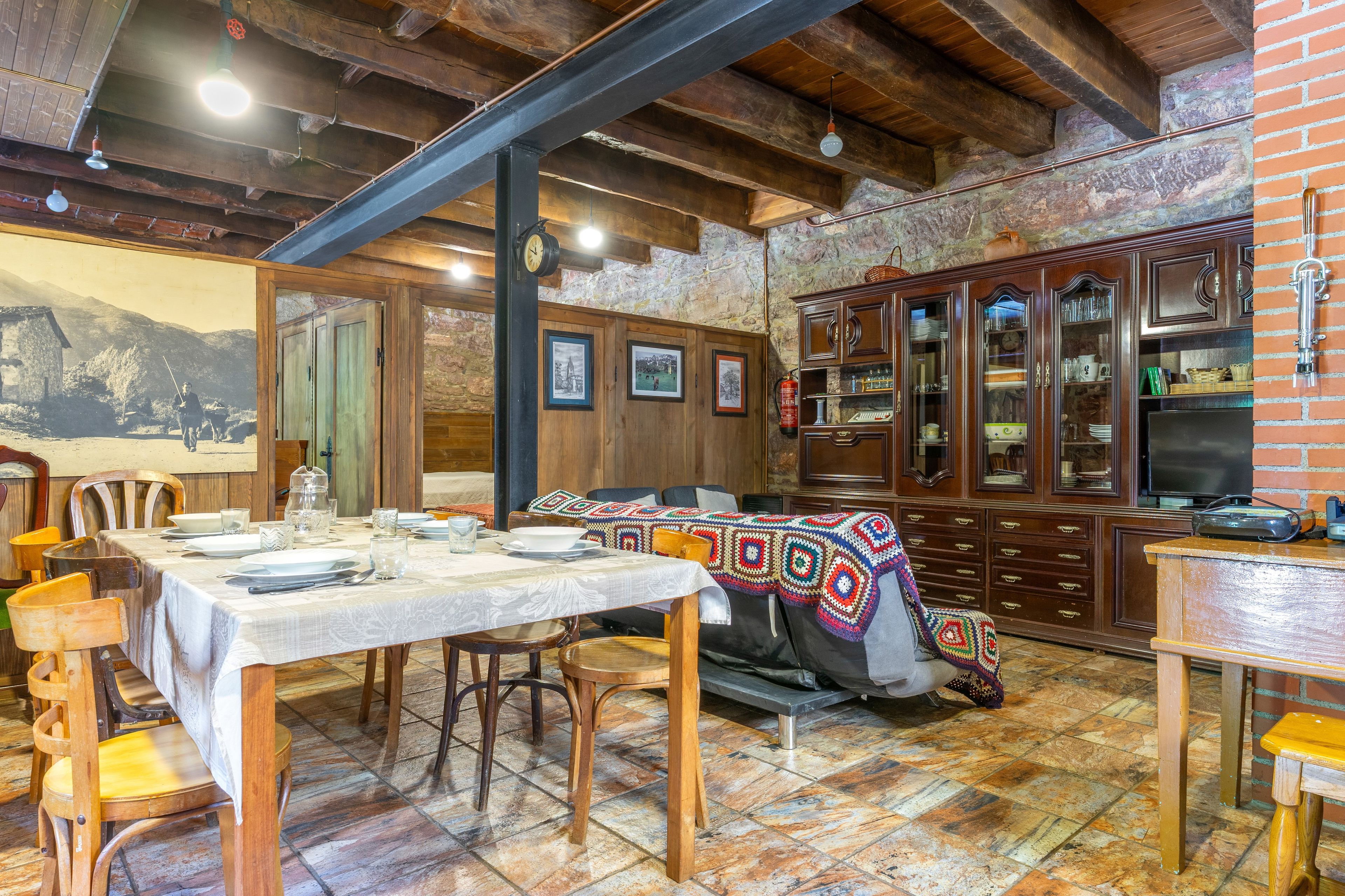 A dining area with a table set for two, wooden chairs, a patterned tablecloth, and a cabinet with glass doors.