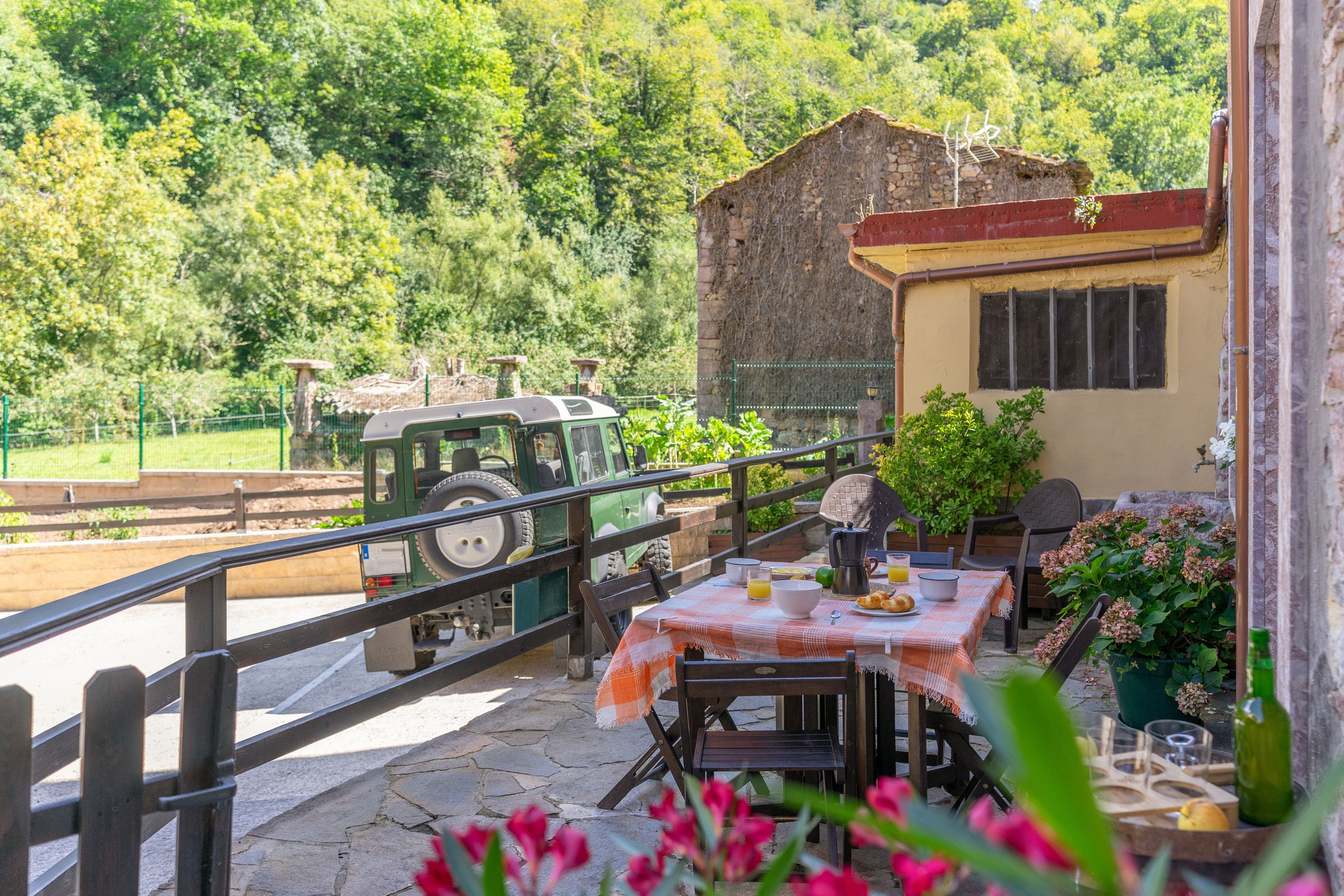A rustic outdoor seating area with a table set for two, overlooking a road with a vintage green vehicle.