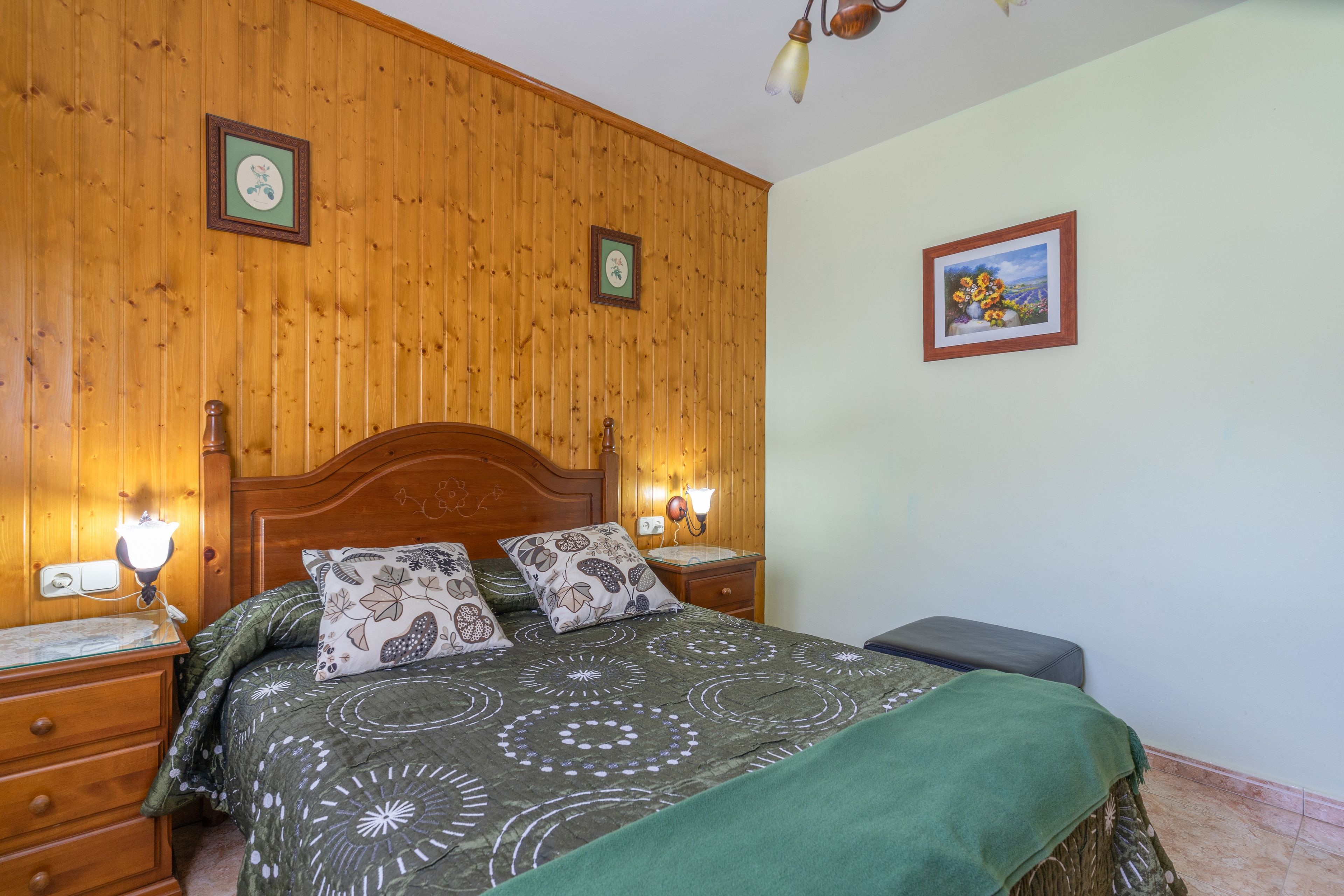 Een slaapkamer met houten lambrisering, een bed, twee bedlampen, een nachtkastje en een lijst met een foto aan de muur.