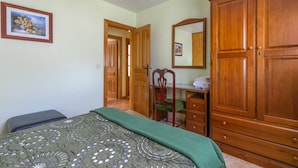 A bedroom with a bed, a wooden wardrobe, a desk with a chair, and a mirror on the wall.