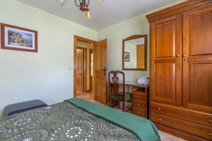 A bedroom with a bed, a wooden wardrobe, a desk with a chair, and a mirror on the wall.