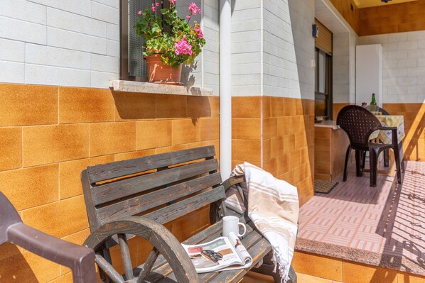A wooden bench with a newspaper and a mug, a potted plant with pink flowers, and a table with chairs outside a building with tiled walls.