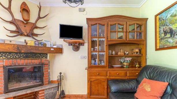 A living room with a fireplace, a television mounted on a wooden stand, a leather sofa, and a cabinet with glass doors.
