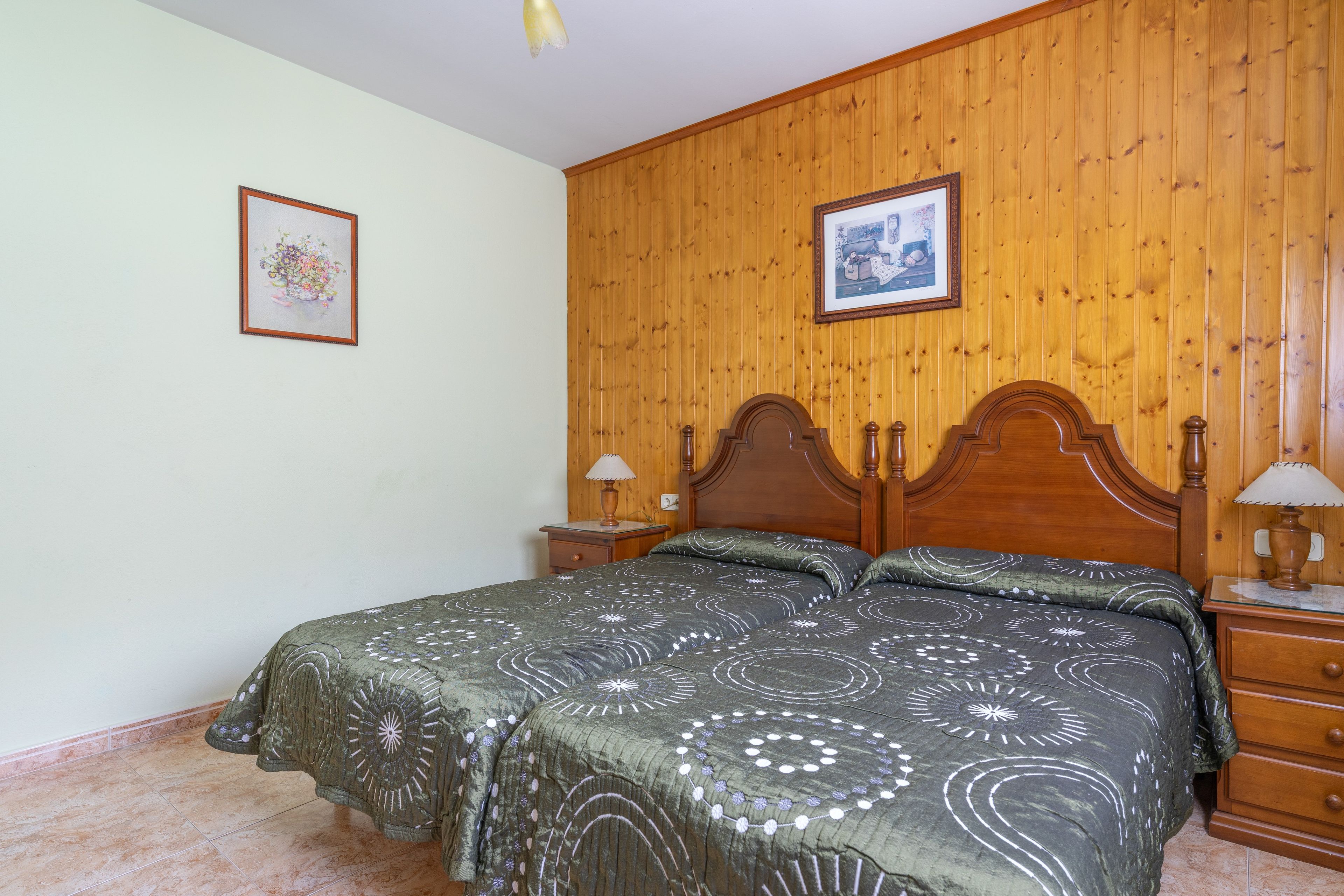Une chambre à coucher avec une tête de lit en bois, deux tables de chevet avec des lampes, un lit recouvert d’une couvre-lit à motifs et deux tableaux encadrés accrochés aux murs.