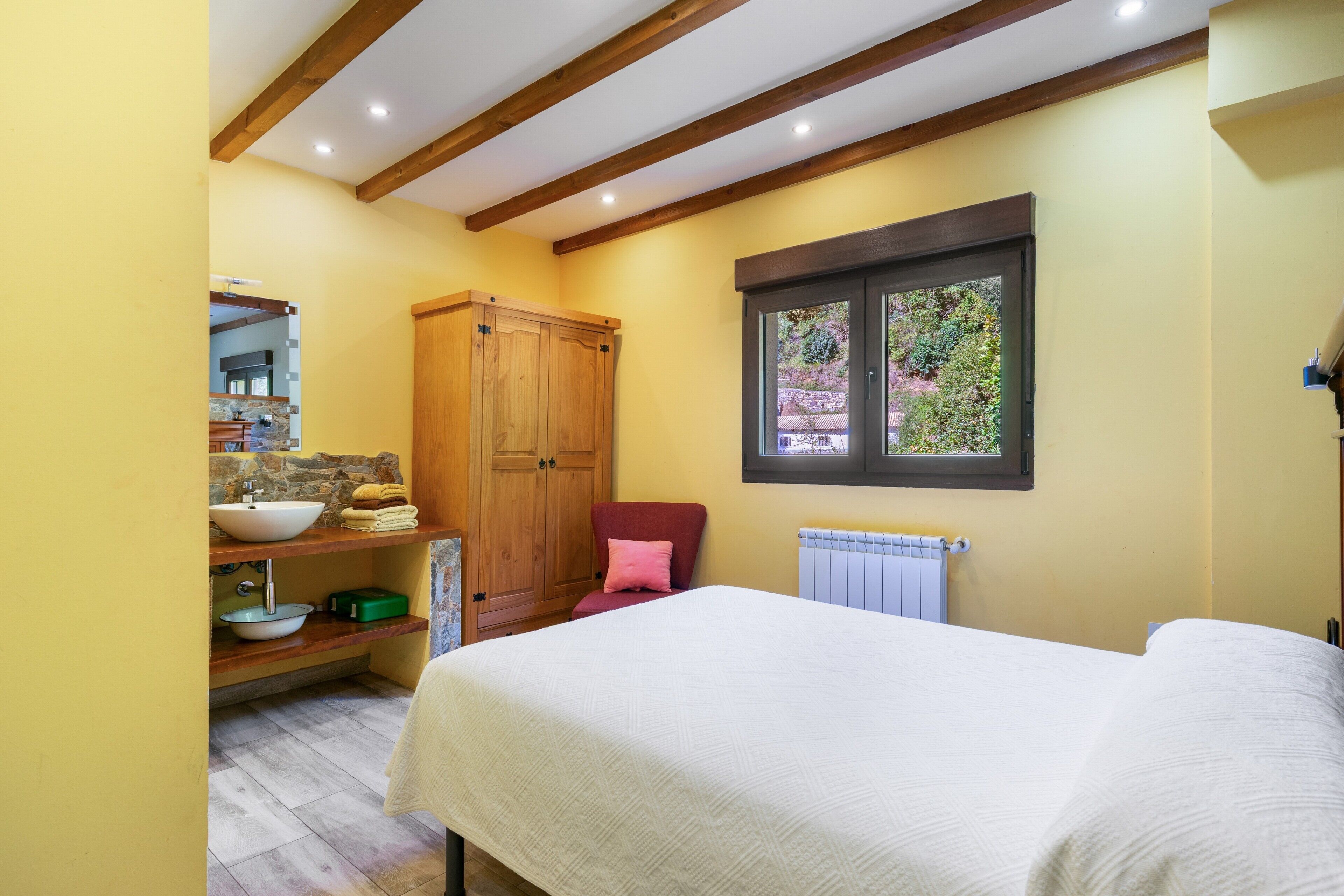 Een slaapkamer met een bed, een houten garderobe, een badkamer met een wastafel en spiegel, en een raam met uitzicht op groen.