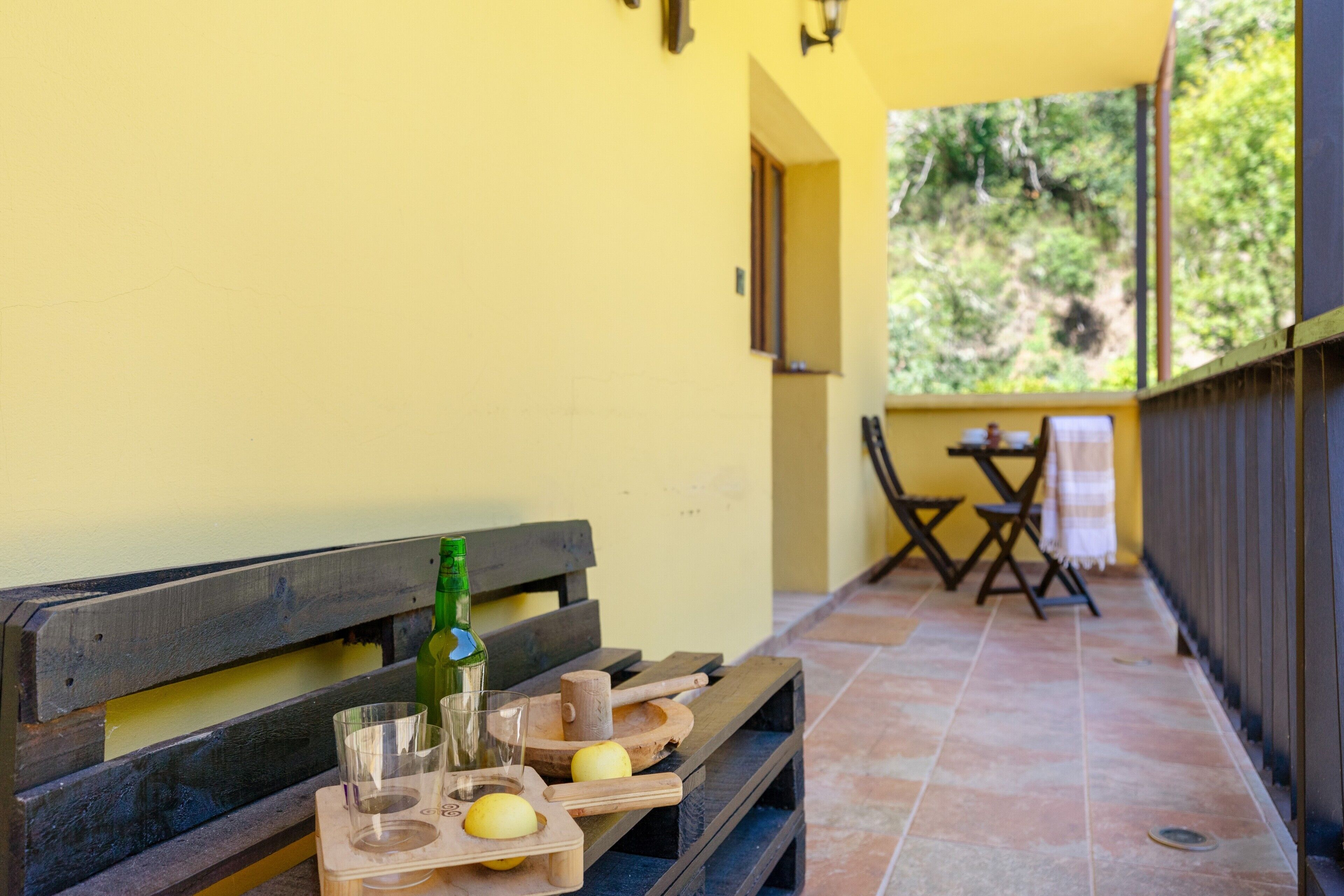 A wooden bench with a bottle and glasses on it, and a table with a cloth in the background.