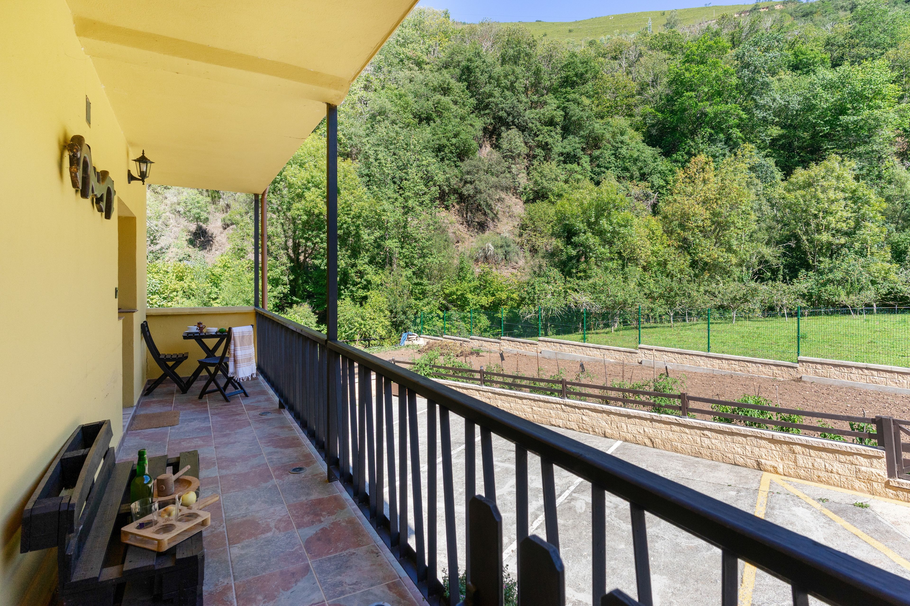Un balcón con pared amarilla, una mesa con sillas y vista a un paisaje verde con árboles y una cerca.
