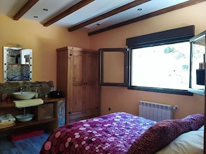A bedroom with a bed, a window with a view, a wooden wardrobe, and a bathroom with a sink and mirror.