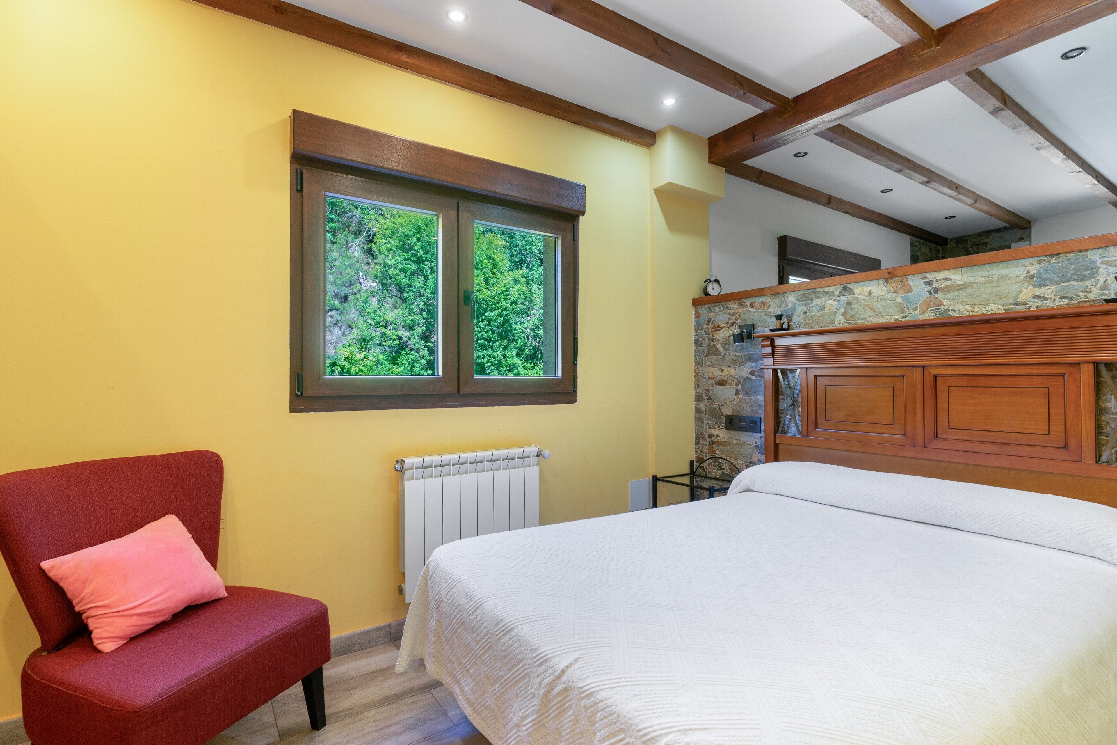 Un dormitorio con una cama, una silla con cojín, una ventana, un radiador y una pared de piedra.