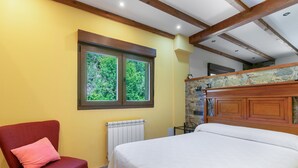 Une chambre à coucher avec un lit, une chaise avec un coussin, une fenêtre, un radiateur et un mur en pierre.