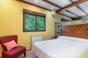 Un dormitorio con una cama, una silla con cojín, una ventana, un radiador y una pared de piedra.