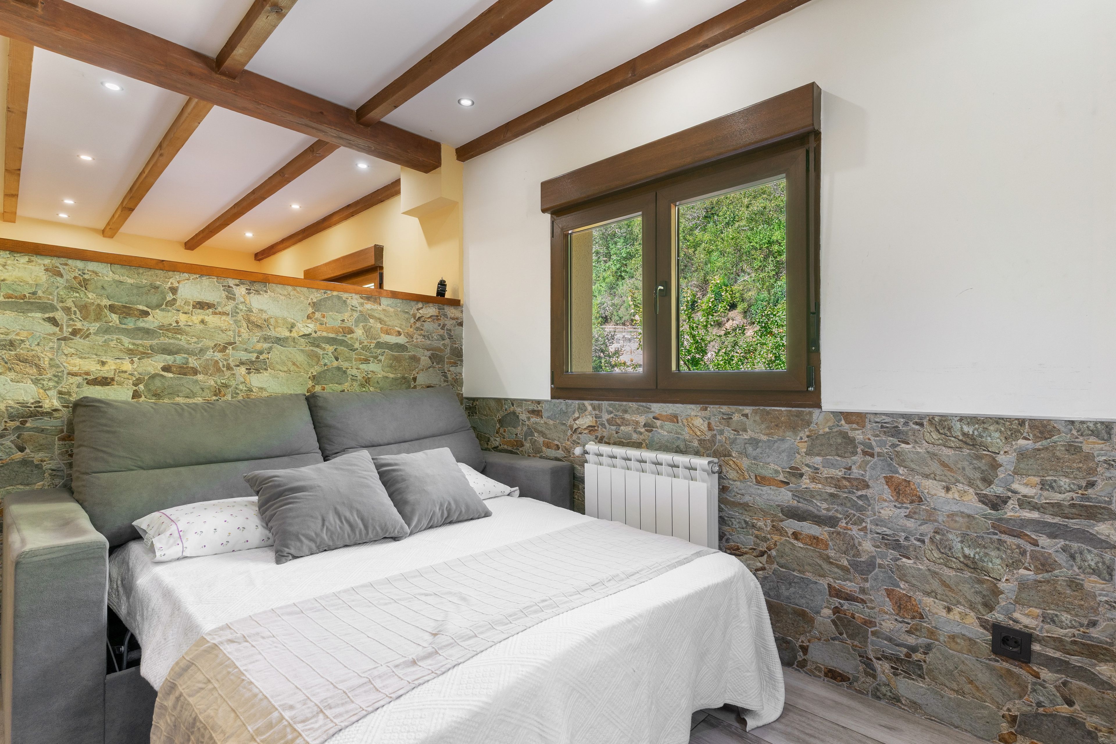 Un dormitorio con pared de piedra, cama con colcha blanca y ventana con vista a follaje.