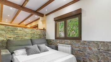 Un dormitorio con pared de piedra, cama con colcha blanca y ventana con vista a follaje.