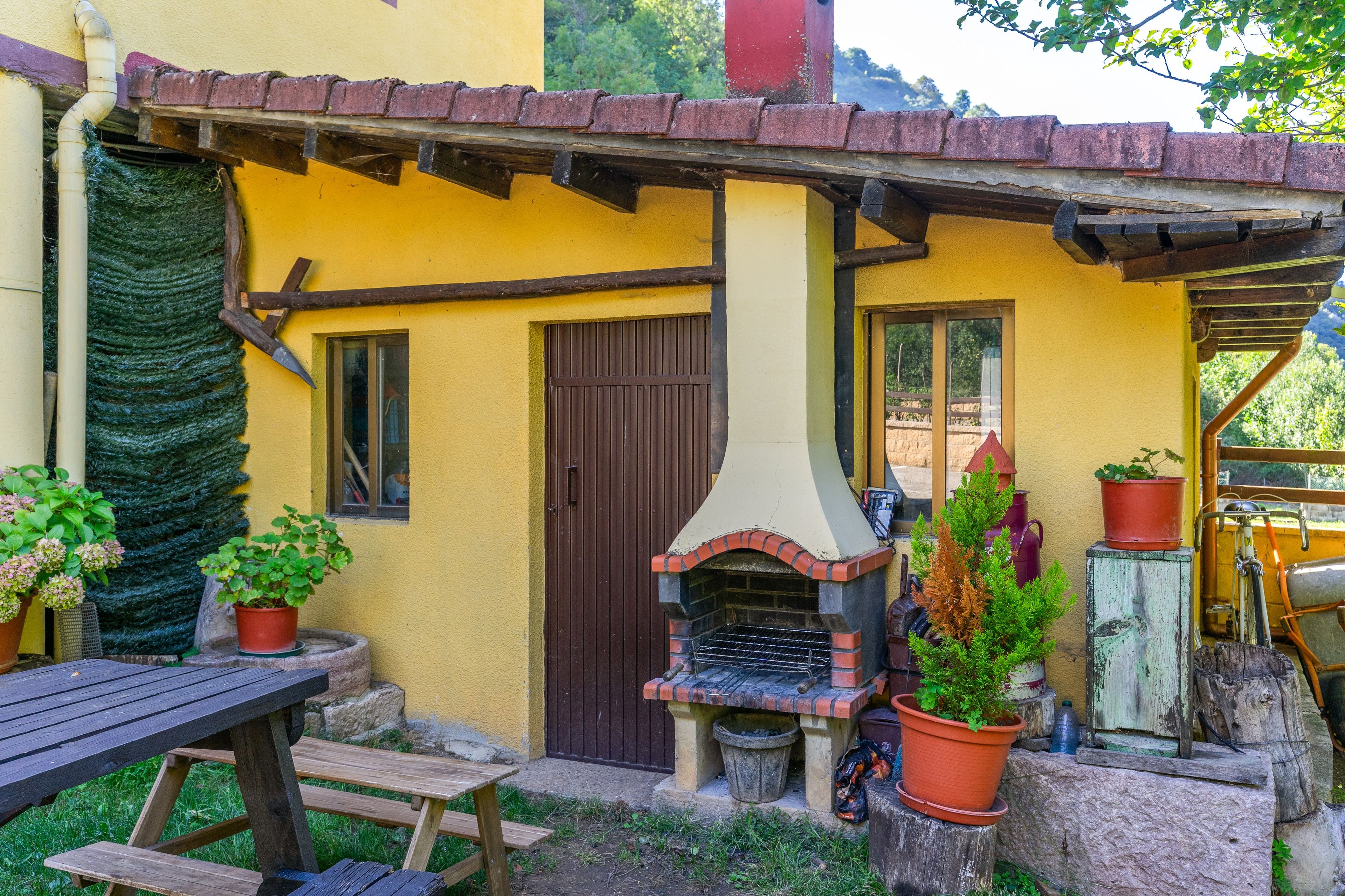 Un área de estar al aire libre de estilo rústico, con una mesa de madera, bancos, un horno de ladrillo y plantas en macetas.