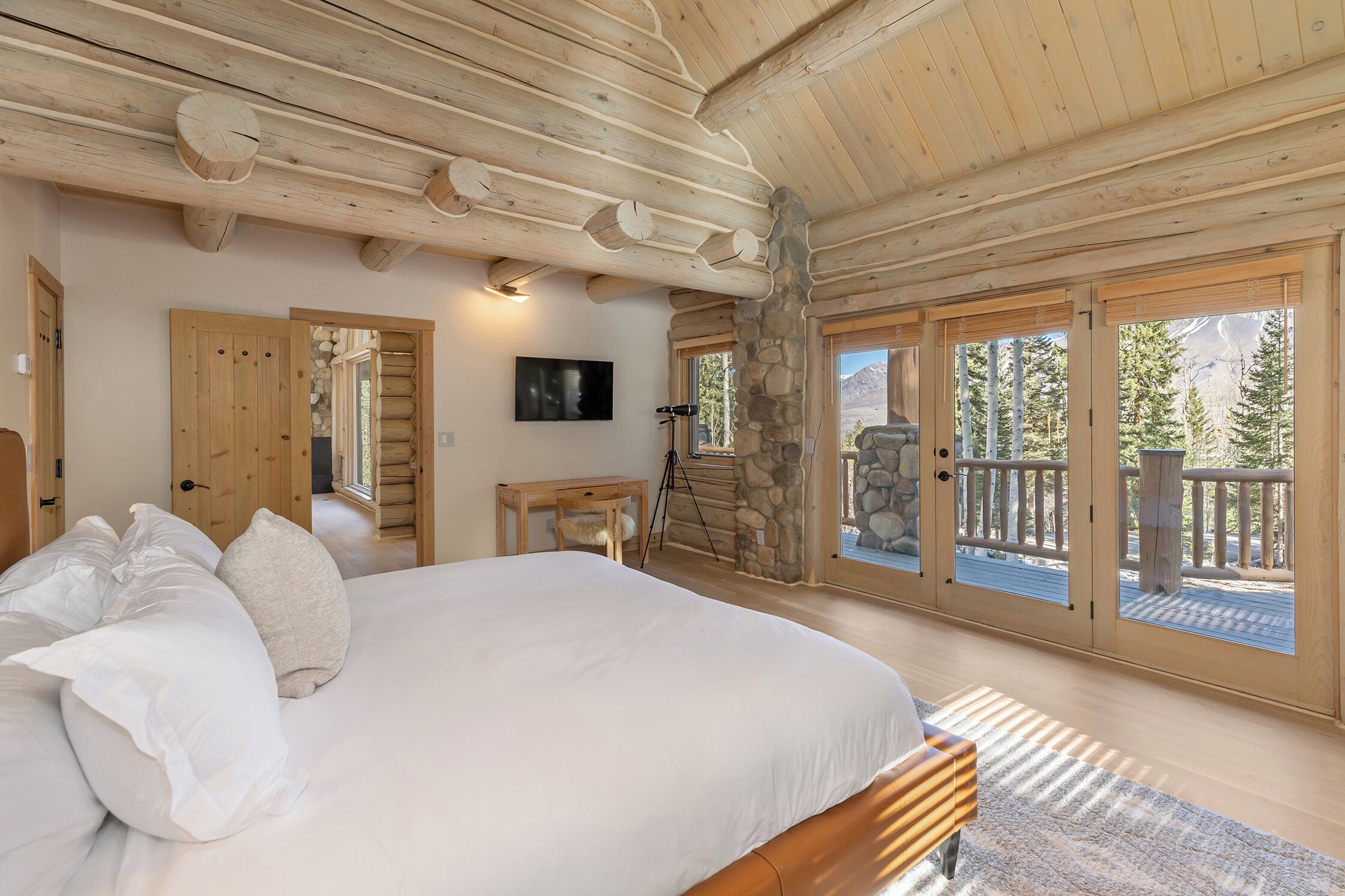 Ein Schlafzimmer mit einem großen Bett, Holzbalken an der Decke, einer Steinwand und einem Balkon mit Blick auf Bäume.