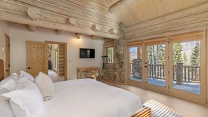 Ein Schlafzimmer mit einem großen Bett, Holzbalken an der Decke, einer Steinwand und einem Balkon mit Blick auf Bäume.