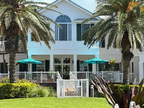 A two-story building with a white exterior, a balcony with a white railing, and a covered outdoor seating area with umbrellas.