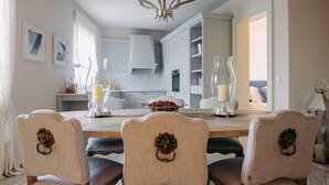 Een eetruimte met een houten tafel, vier gestoffeerde stoelen met decoratieve rugleuningen, een kroonluchter, en een keuken op de achtergrond.