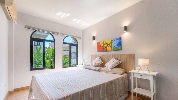Une chambre avec un grand lit, une table de chevet, une lampe fixée au mur et une fenêtre donnant sur un paysage verdoyant.