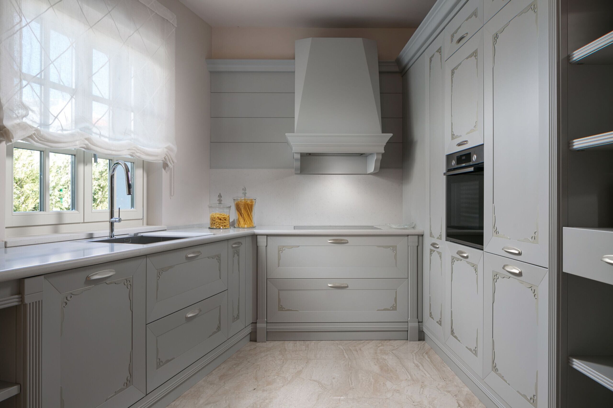 Una cocina con muebles blancos, una ventana con cortinas y un horno empotrado.