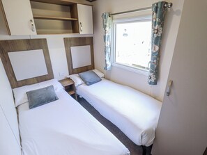 A small, compact room with two single beds, a wooden headboard, a window with floral curtains, and a built-in shelf above the beds. - 39 Larch View With hot tub (Lincoln)