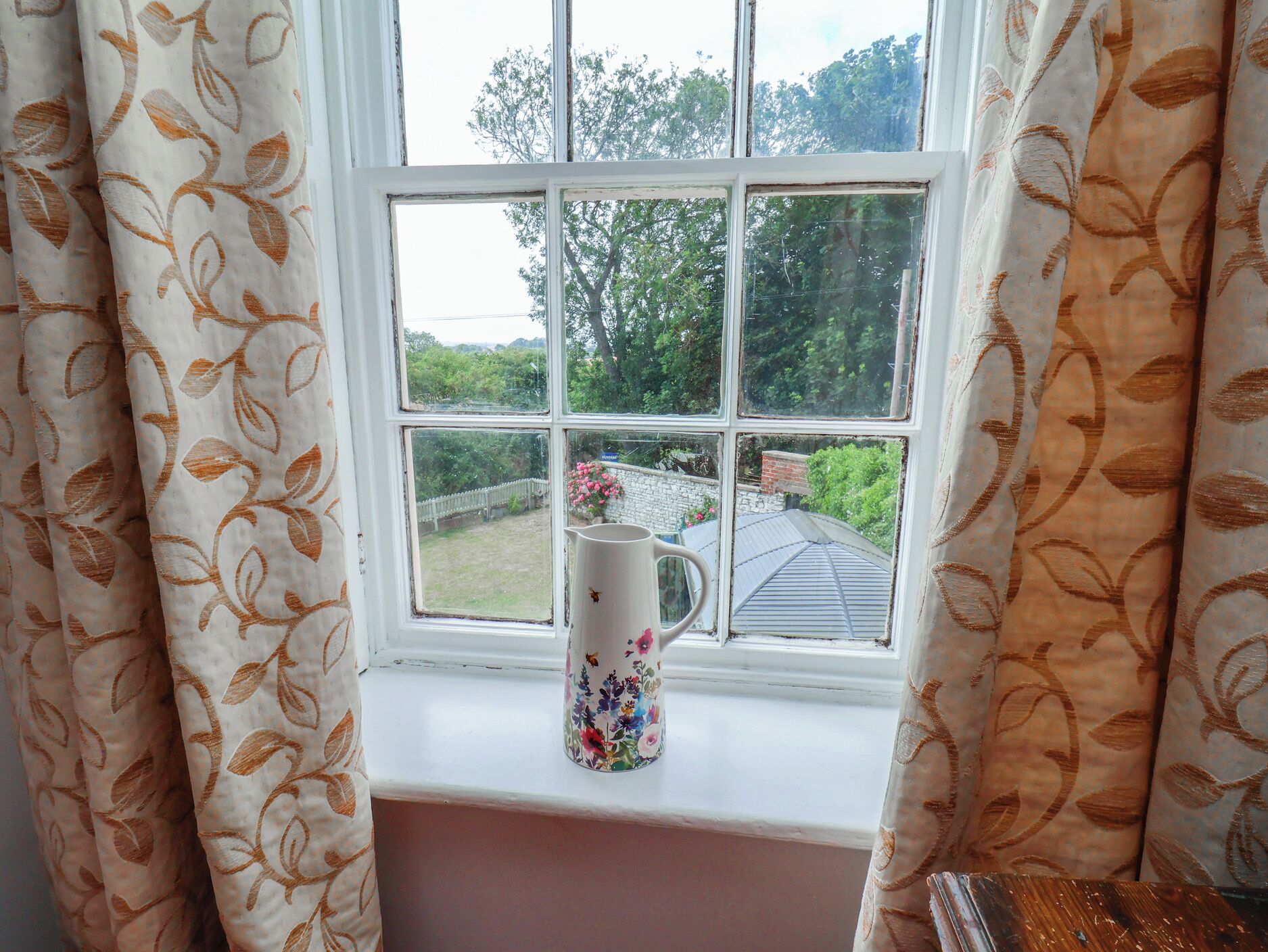 A window with a floral-patterned curtain, a white pitcher with a floral design, and a view of a garden and trees outside.