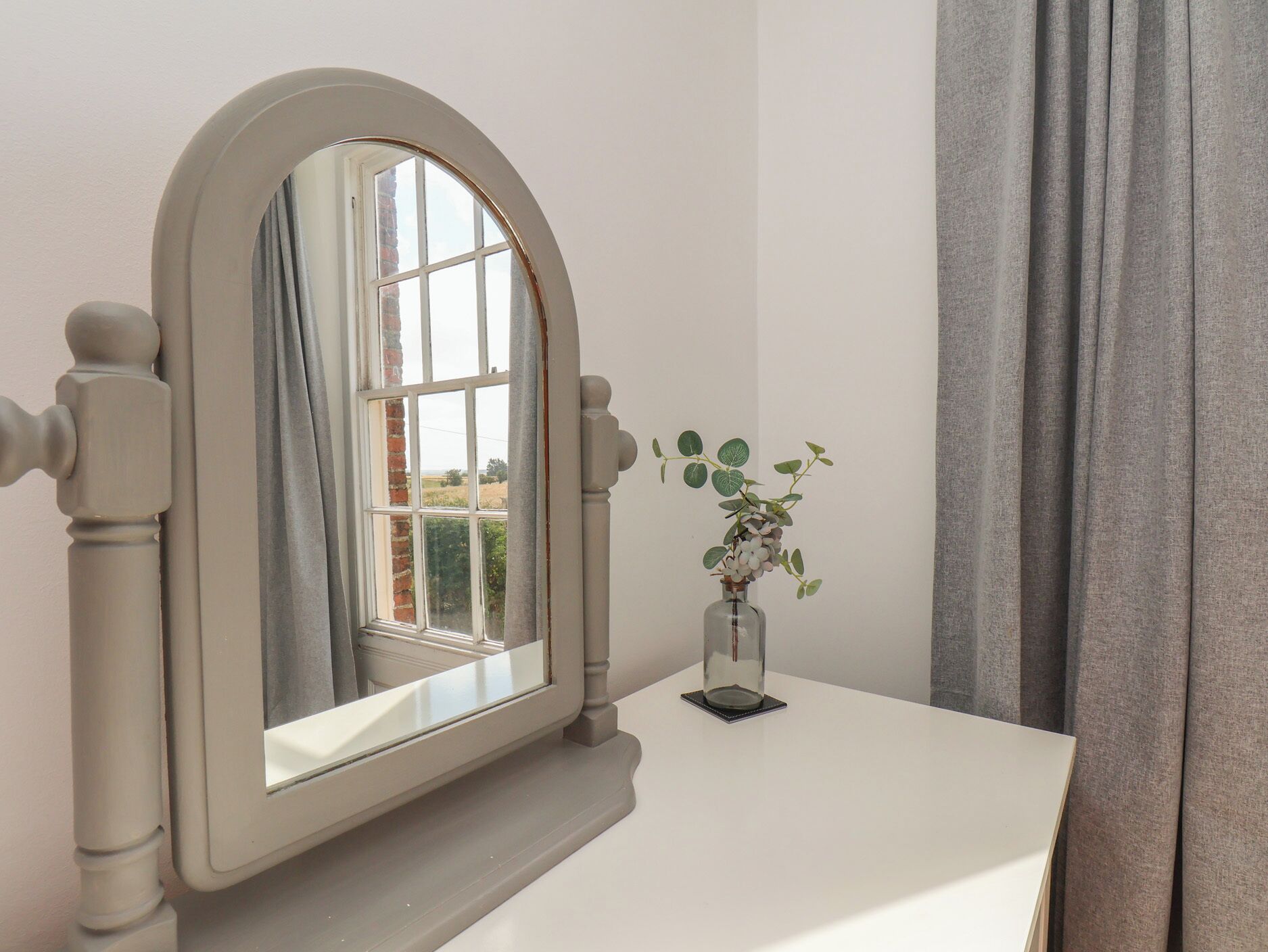 Ein Zimmer mit weißem Schminktisch, einem Spiegel mit gebogenem Rahmen, einem Fenster mit Blick ins Grüne und einer Vase mit Pflanze.