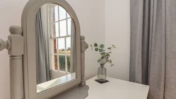 Ein Zimmer mit weißem Schminktisch, einem Spiegel mit gebogenem Rahmen, einem Fenster mit Blick ins Grüne und einer Vase mit Pflanze.
