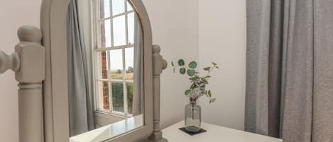 Una habitación con un tocador blanco, un espejo de marco arqueado, una ventana con vistas a follaje y un jarrón con una planta.