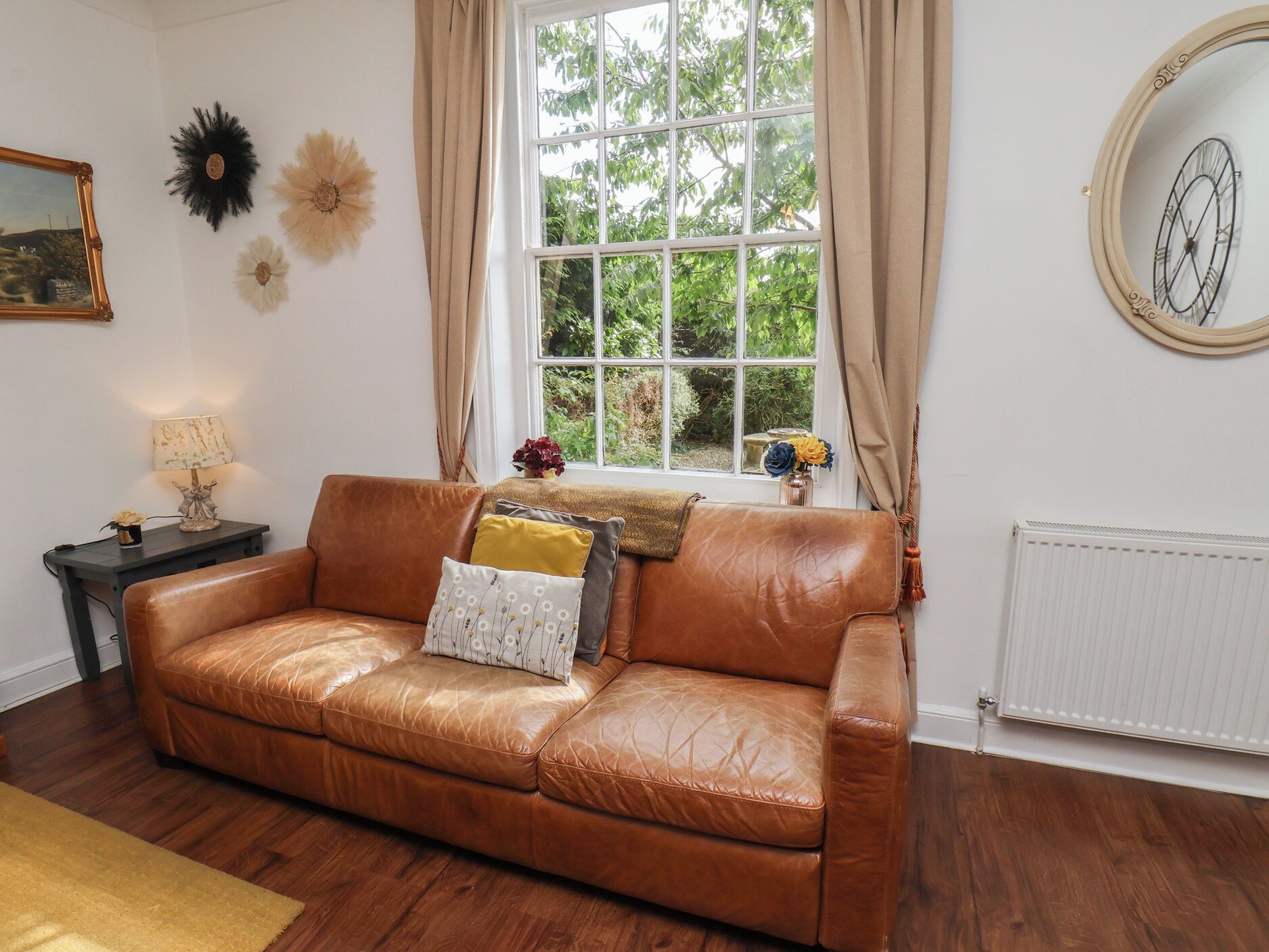 A brown leather sofa with cushions, a small table with a lamp, and a window with curtains.