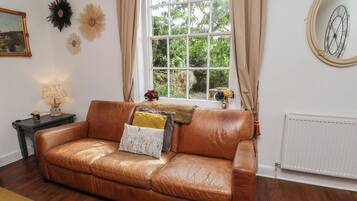 A brown leather sofa with cushions, a small table with a lamp, and a window with curtains.