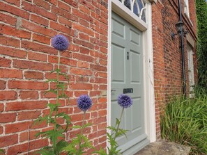 A brick building with a white door and a small garden area with purple flowers. - Reighton House (Filey)
