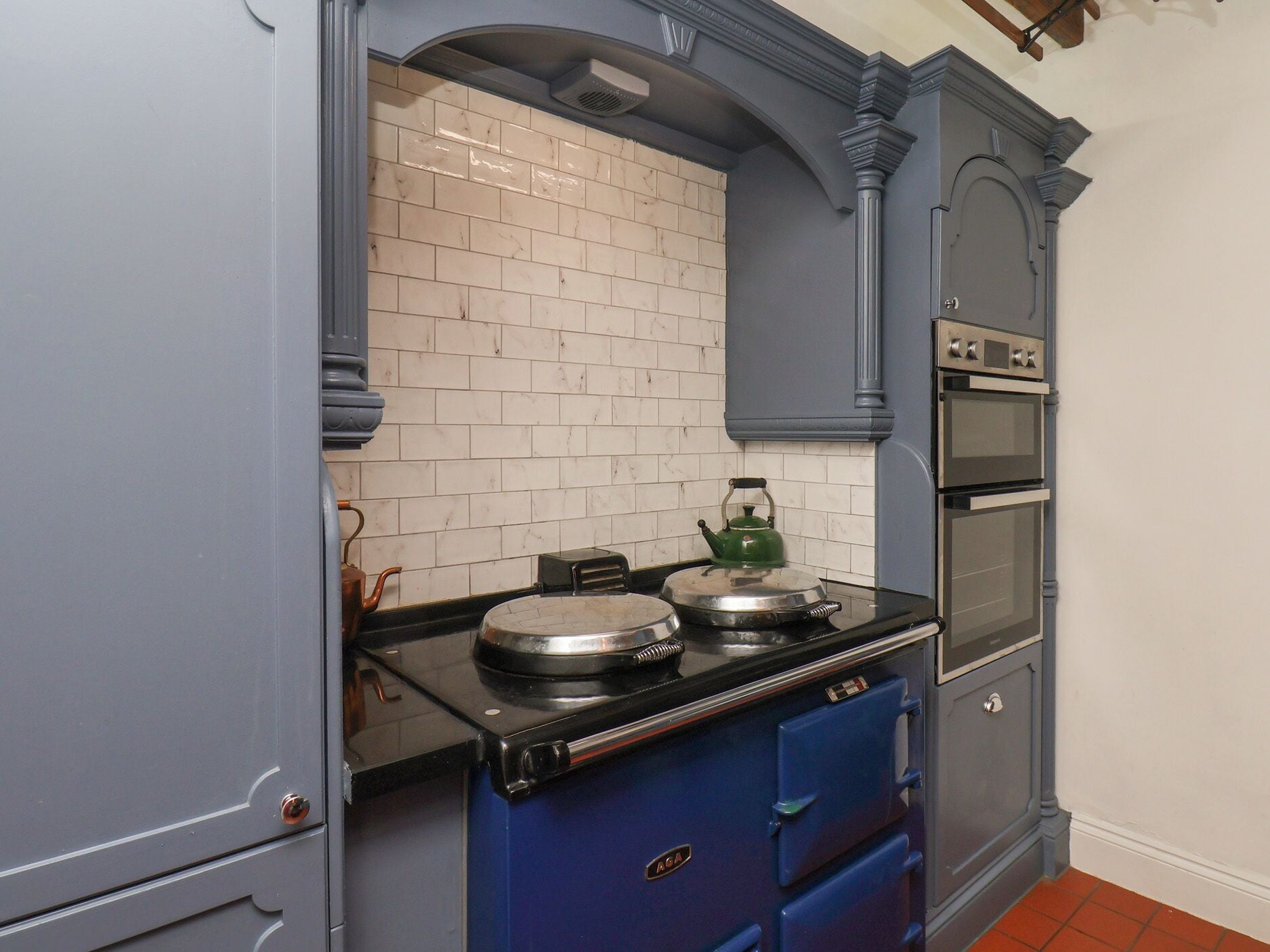 Una cocina con estufa negra, hervidor verde y salpicadero de azulejos blancos.