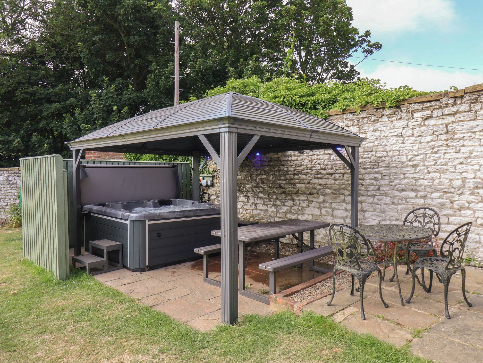 A covered outdoor seating area with a hot tub, picnic tables, and metal chairs.
