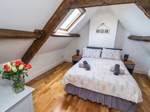 An attic bedroom with a sloped ceiling, a bed with a patterned bedspread, two bedside tables with lamps, and a vase of red and white roses on a dresser. - Reighton House (Filey)