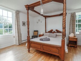 Una cama con dosel de madera y un colgante, una mesita de noche con un adorno, y una ventana con cortinas.