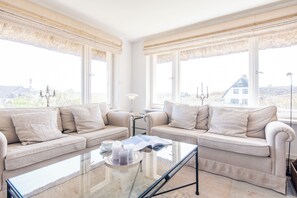 Una habitación con dos sofás, una mesa de centro de vidrio y ventanas amplias con cortinas.