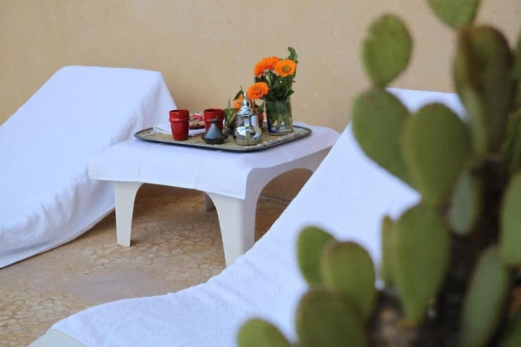 A sunlit outdoor seating area with a table set for two, featuring a tray with a vase of orange flowers and a small bell.