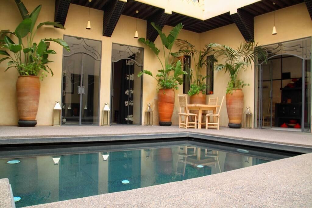 Un área de piscina con una mesa y sillas, rodeada de grandes plantas en macetas.