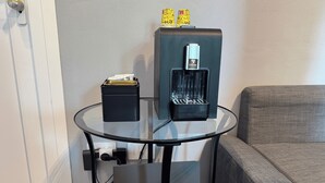 Una mesa de vidrio con una cafetera y un cesto de basura, un sofá gris, y una puerta al fondo.