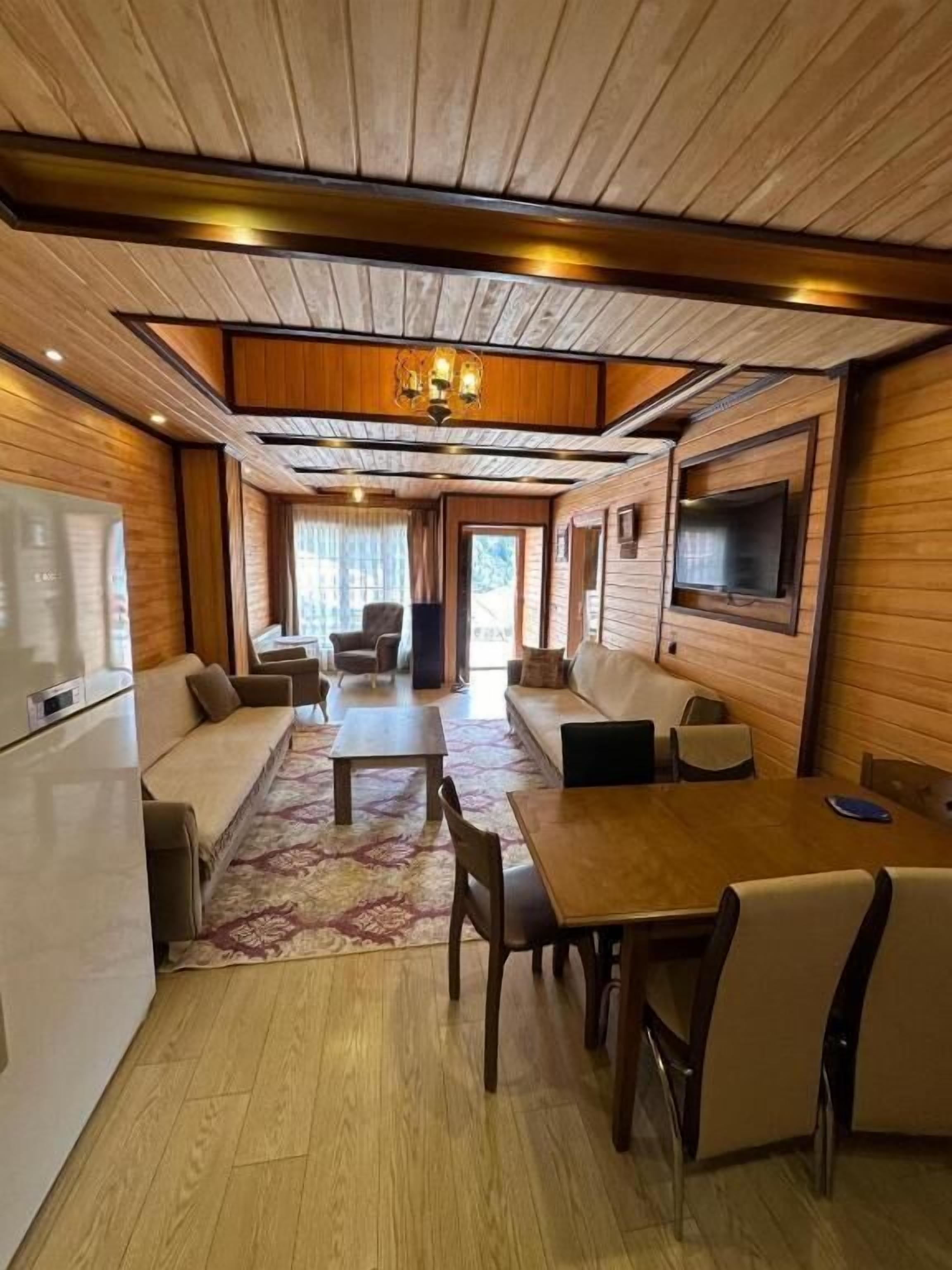 Una habitación con techo de madera, una mesa de comedor con sillas, un sofá, un televisor montado en la pared y un refrigerador.