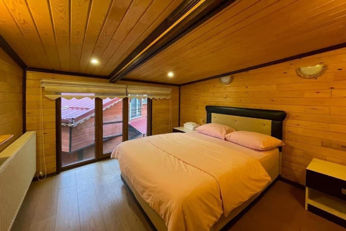 A bedroom with a wooden ceiling, a bed with pink bedding, a nightstand, and a window with a view.