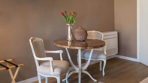 Una habitación con una mesa redonda, dos sillas blancas, un jarrón con flores rojas y un radiador blanco con textura.