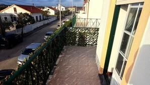 Un balcon doté d’un sol carrelé à motifs, d’une clôture verte et offrant une vue sur une rue où sont garées des voitures.