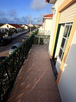 A balcony with a patterned tile floor, a green fence, and a view of a street with parked cars. - Vio's Home (Serra d' El-Rei)